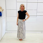 Woman wearing a black top and patterned skirt standing in a room with white walls and a shelf.