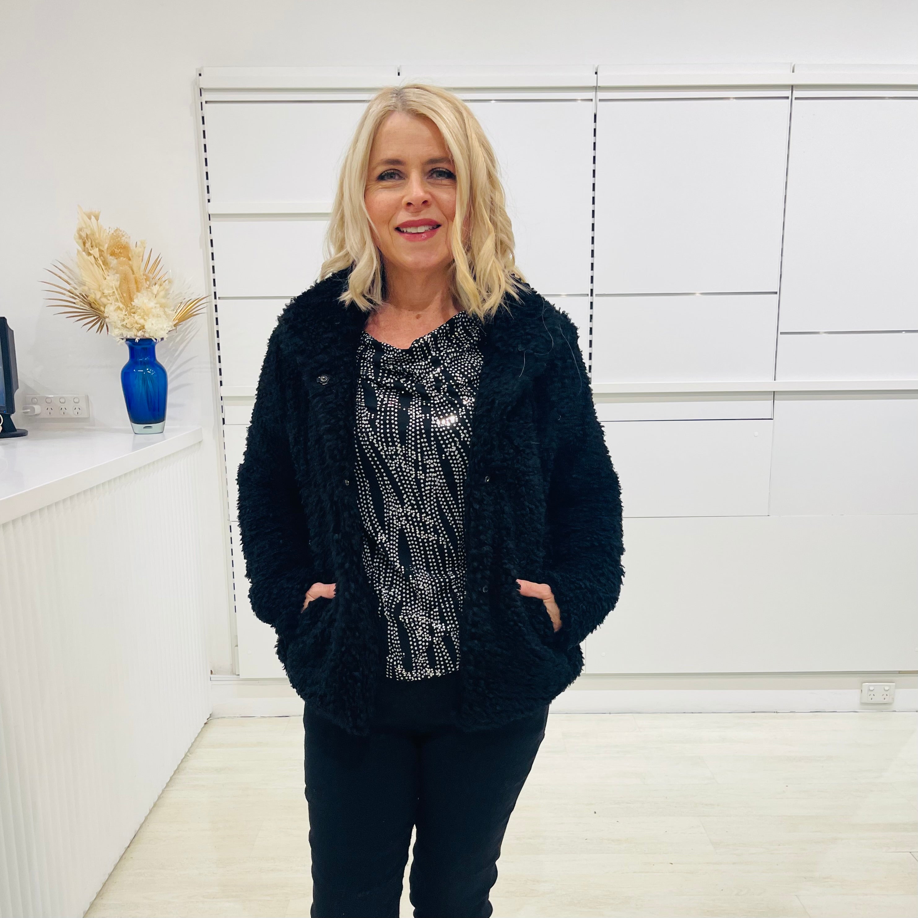 Black fluffy, faux fur, teddy bear jacket with pockets. Worn with black tailored fitted pants and a black and silver sparkly cowl neckline top.
