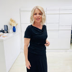 Woman in a black dress standing in a room with white walls and a desk.