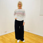 Woman wearing a striped sweater and black pants standing in a room with white walls and wooden floor.