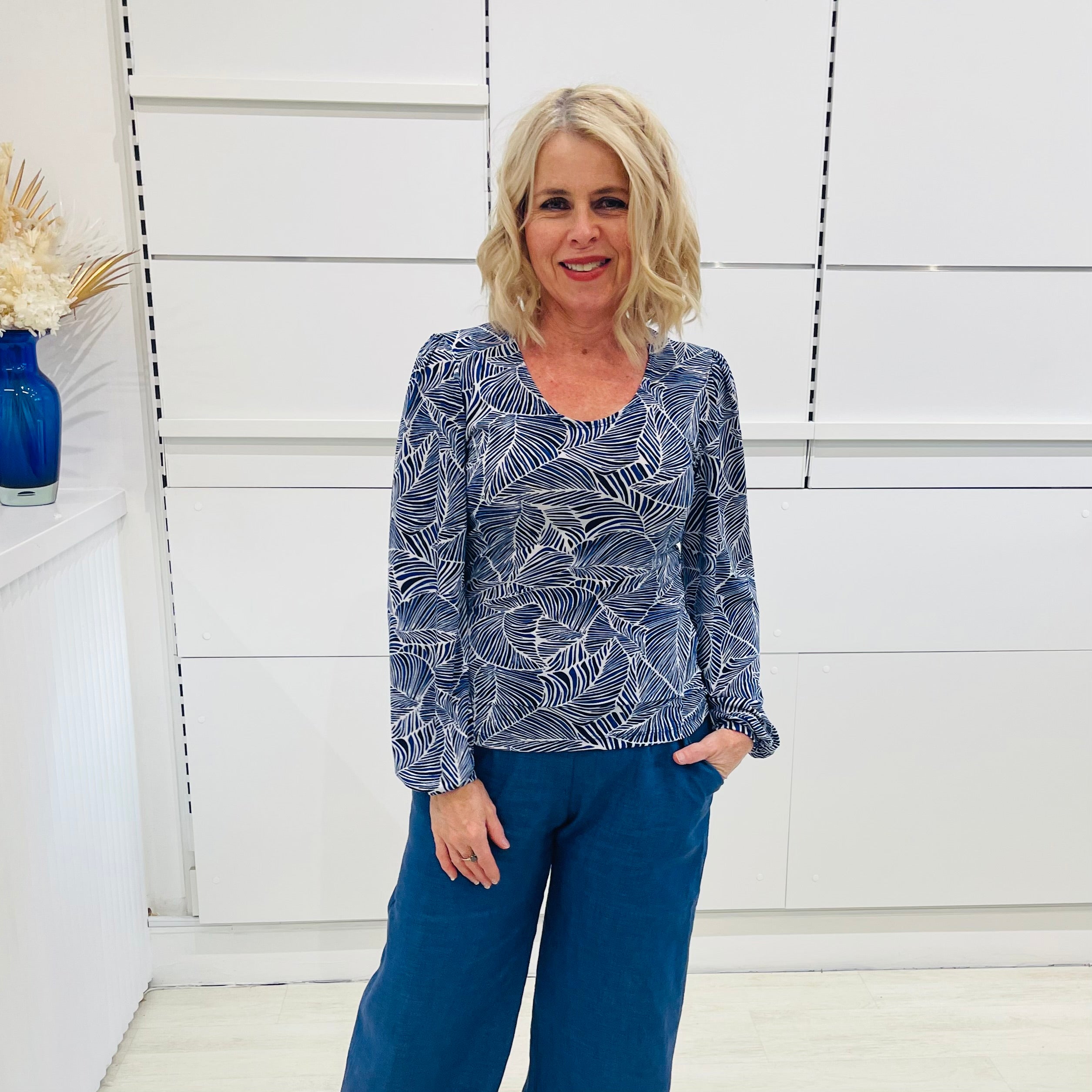 Woman wearing a long sleeve, semi fitted, flowy blue top with a delicate leaf print, paired with denim blue colored linen pants.