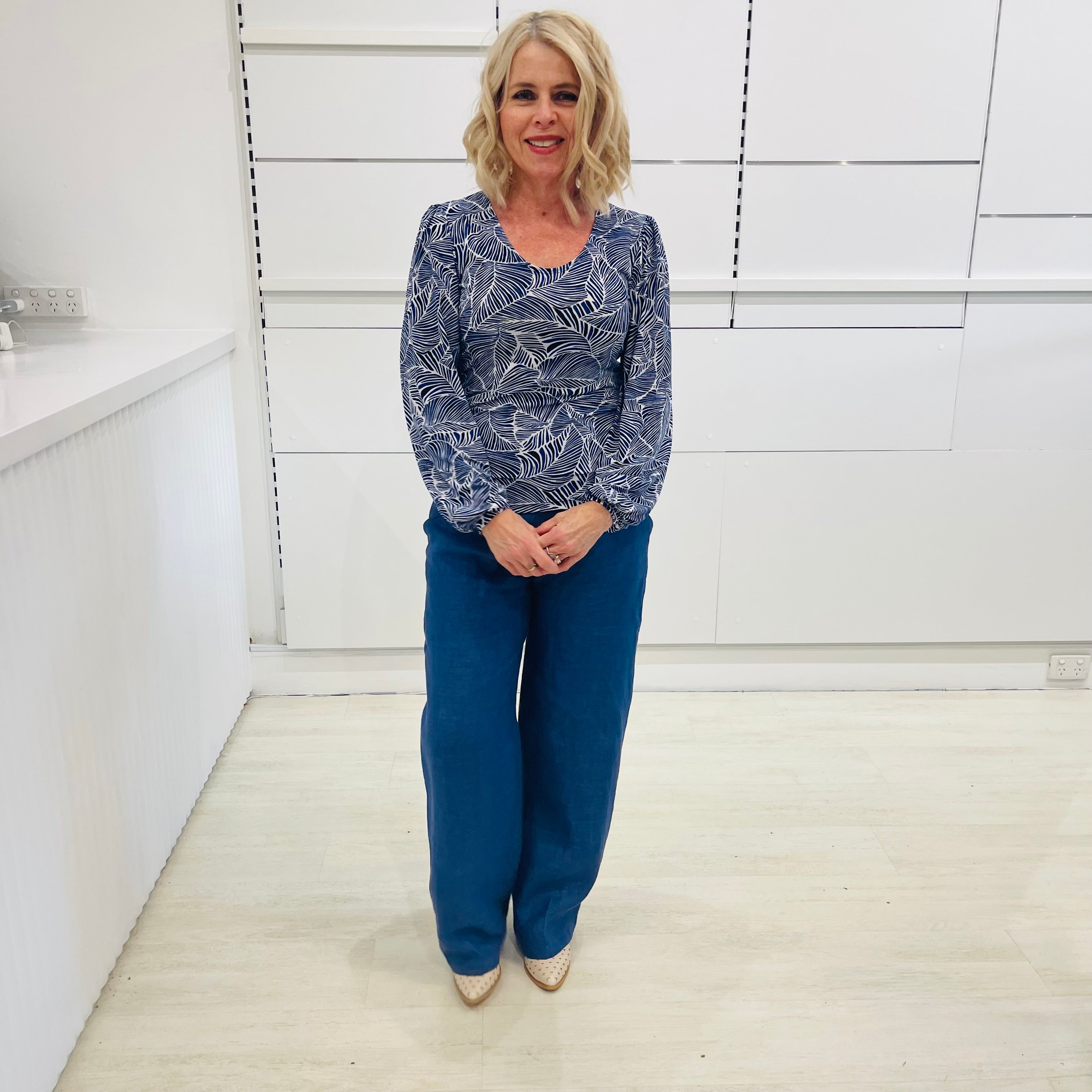 Woman wearing a long sleeve, semi fitted, flowy blue top with a delicate leaf print, paired with denim blue colored linen pants.