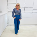 Woman wearing a long sleeve, semi fitted, flowy blue top with a delicate leaf print, paired with denim blue colored linen pants.