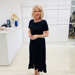 Woman in a black dress standing in a room with white walls and a desk.