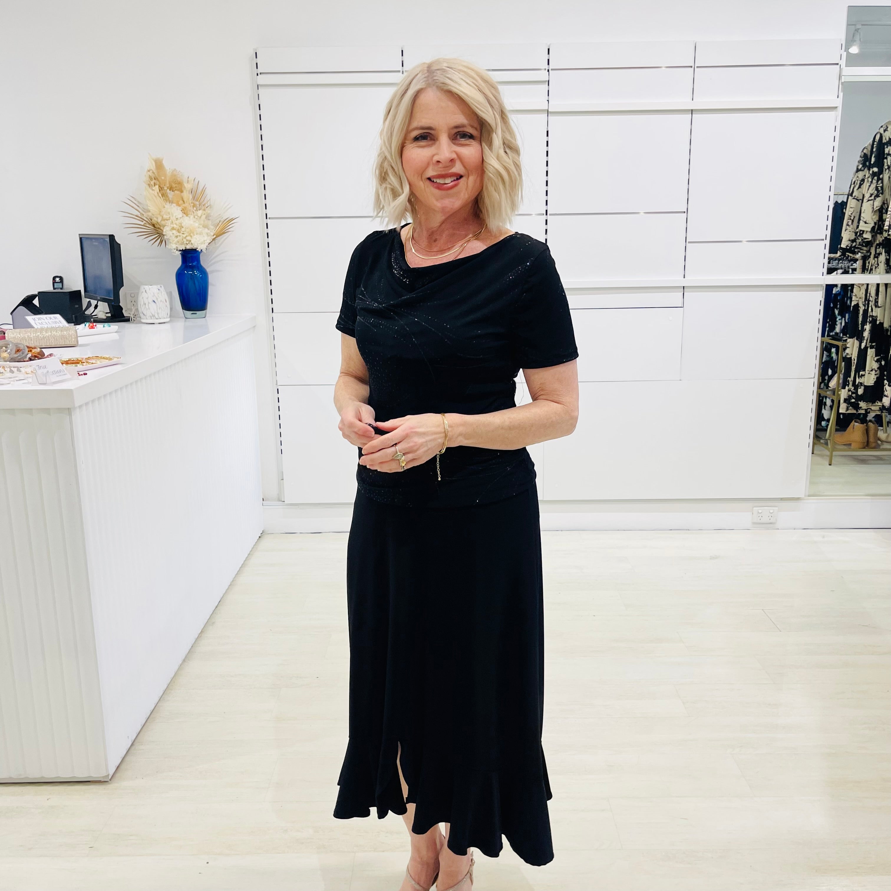 Woman in a black dress standing in a room with white walls and a desk.