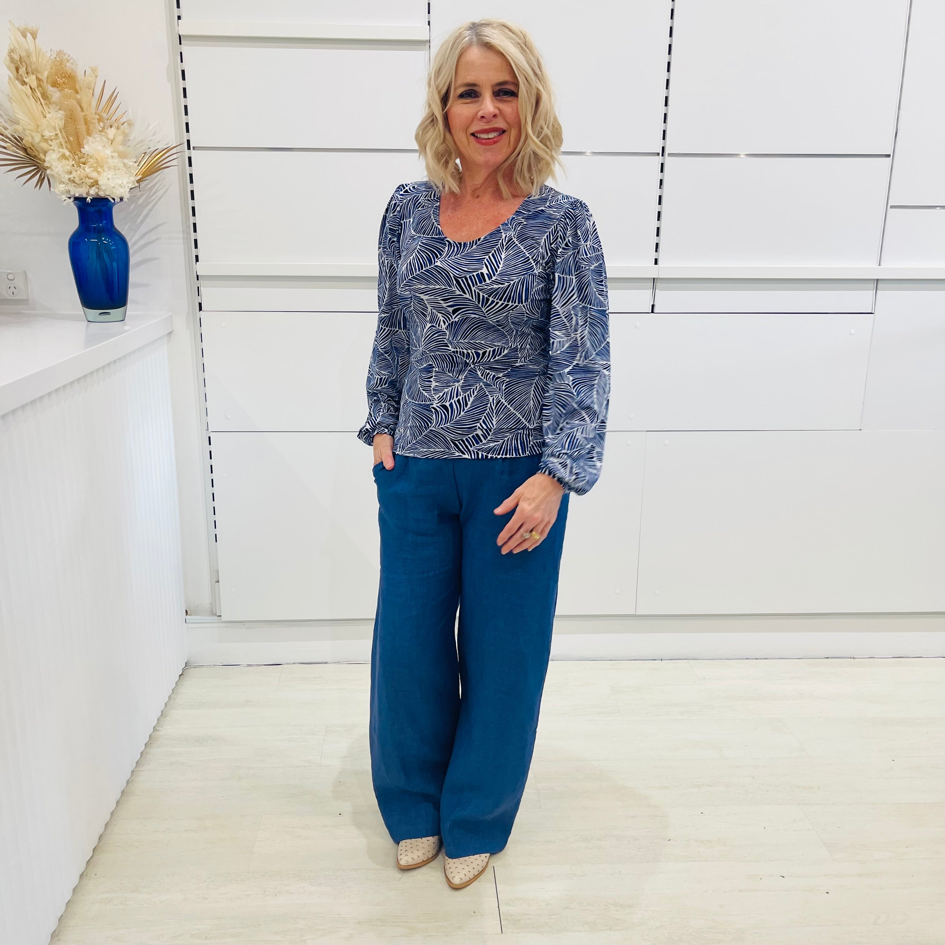 Woman wearing a flowy blue top with long sleeves, a delicate leaf print, paired with denim blue colored linen pants.