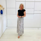 Woman in a black top and patterned skirt standing in a room with white walls and a vase on a shelf.