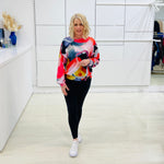 Woman wearing a colorful sweater and black leggings in a room with white walls and a shelf.