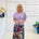 Woman wearing a light purple top and colorful skirt standing in a room with white walls and decorative elements.