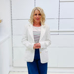 Woman wearing a white blazer over a striped shirt with blue pants against a white wall.