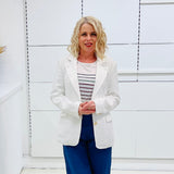Woman wearing a white blazer over a striped shirt with blue pants against a white wall.