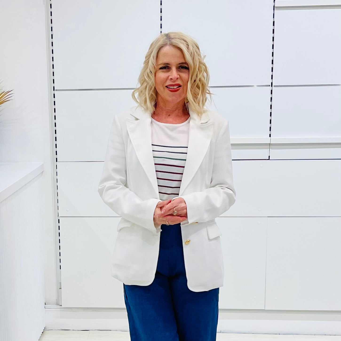 Woman wearing a white blazer over a striped shirt with blue pants against a white wall.