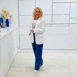 Woman in a white blazer and blue pants standing in a modern interior setting.