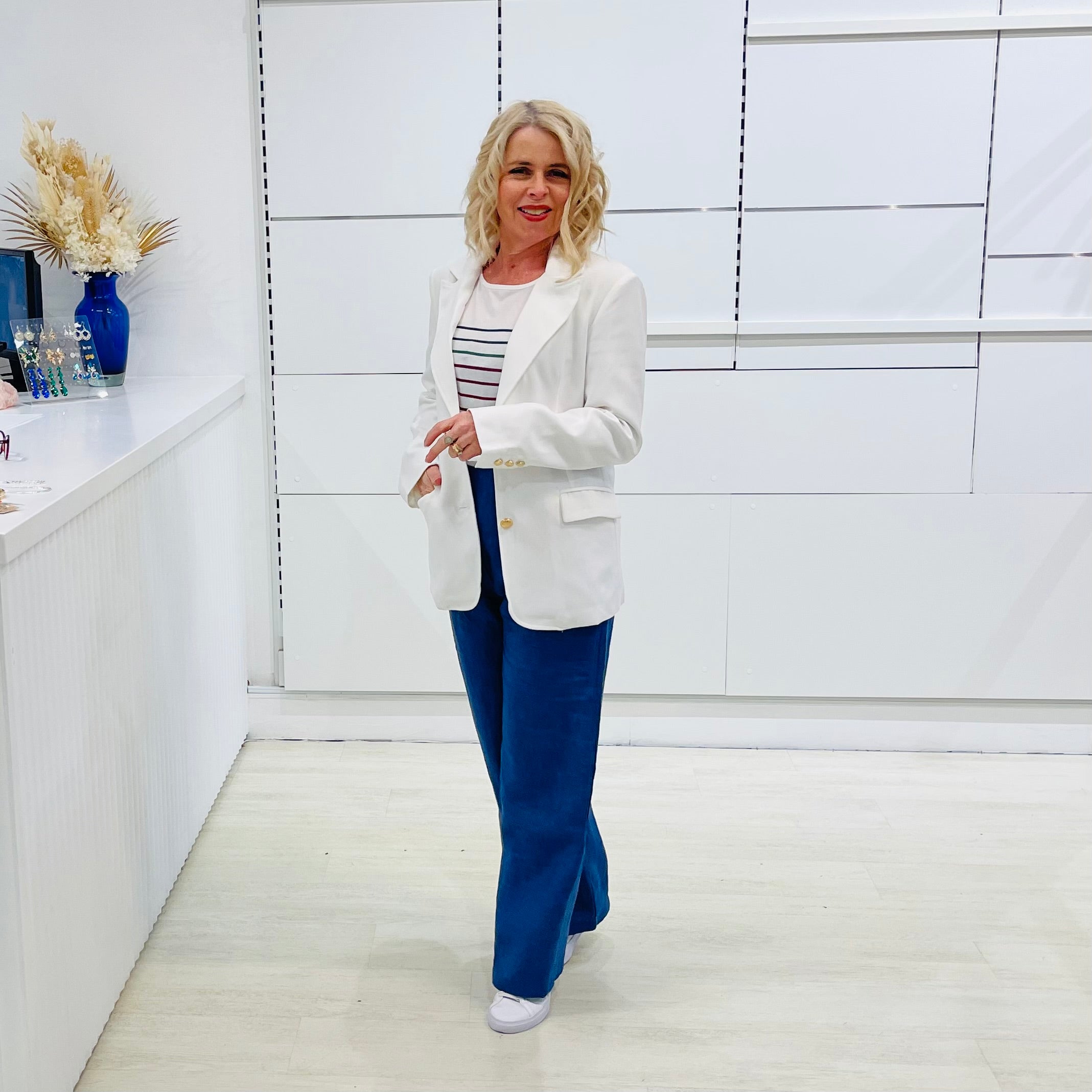 Woman in a white blazer and blue pants standing in a modern interior setting.
