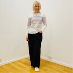 Woman wearing a striped sweater and black pants standing in a room with white walls and wooden floor.