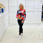 Woman wearing a colorful sweater and black leggings in a white room.