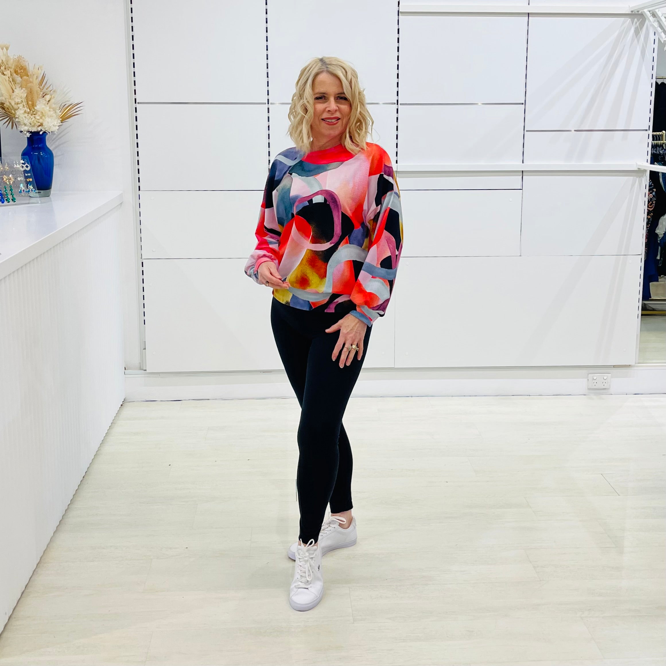 Woman wearing a colorful sweater and black leggings in a white room.
