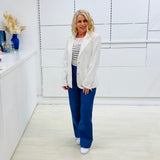 Woman in a white blazer and blue pants standing in a modern interior setting.