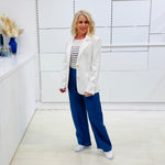 Woman wearing a white blazer, striped shirt, and blue pants in a minimalistic room.