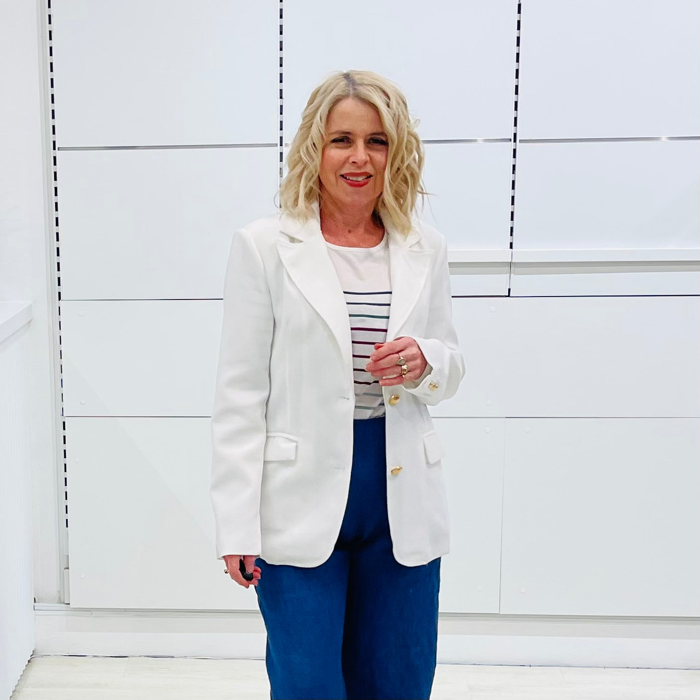 Woman wearing a white blazer over a striped shirt with blue pants against a white background