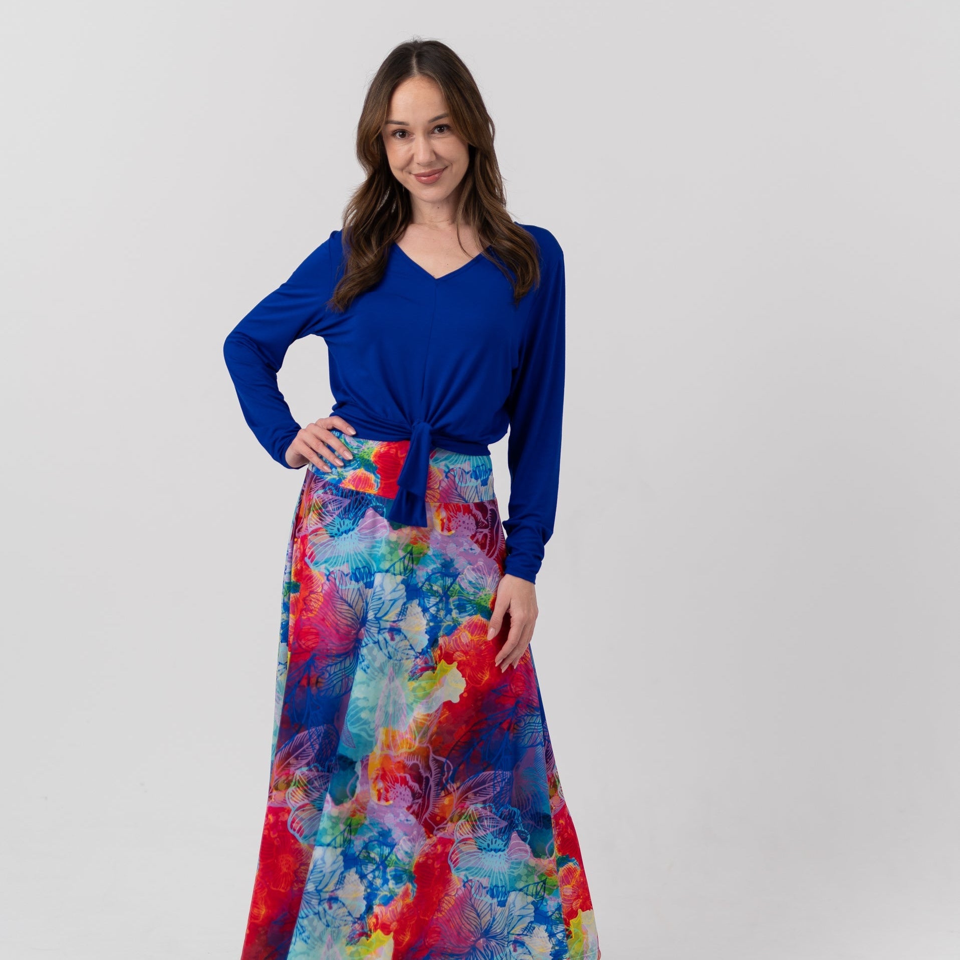 A woman modeling a long-sleeve wrap-style maxi dress with a vibrant blue V-neck bodice and a multicolor abstract watercolor print skirt, standing against a light gray studio background.