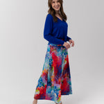 A woman modeling a vibrant, long-sleeved maxi skirt with a colorful abstract watercolor print in shades of blue, red, yellow, and purple, paired with a solid royal blue long-sleeve top, against a light gray studio background.