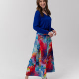 A woman modeling a vibrant, long-sleeved maxi skirt with a colorful abstract watercolor print in shades of blue, red, yellow, and purple, paired with a solid royal blue long-sleeve top, against a light gray studio background.