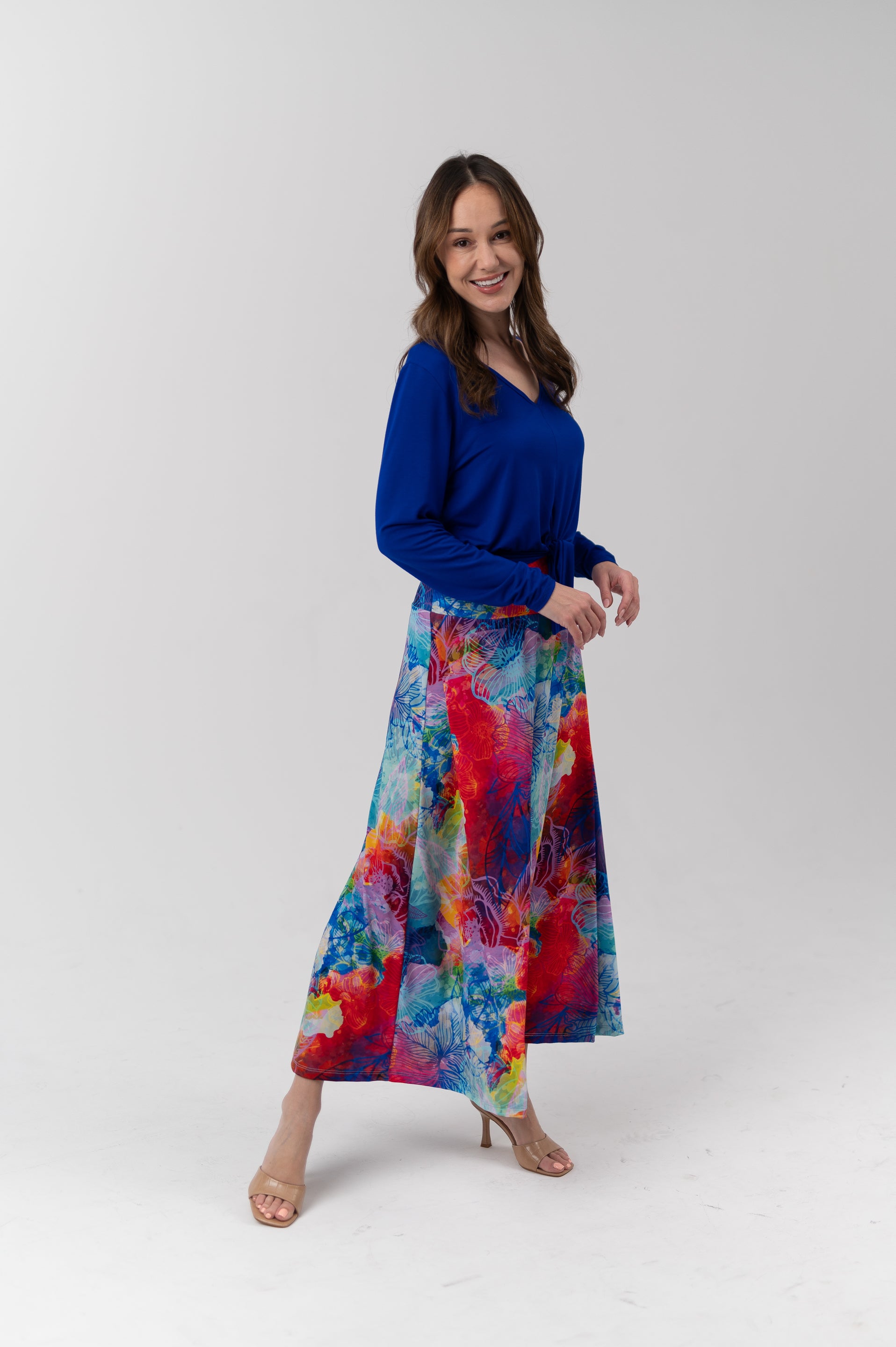 A woman modeling a vibrant, long-sleeved maxi skirt with a colorful abstract watercolor print in shades of blue, red, yellow, and purple, paired with a solid royal blue long-sleeve top, against a light gray studio background.