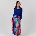 A woman modeling a vibrant, long-sleeved maxi skirt with a colorful abstract watercolor print in shades of blue, pink, red, and yellow, paired with a solid royal blue long-sleeve top, against a light gray studio background.