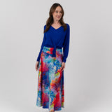 A woman modeling a vibrant, long-sleeved maxi skirt with a colorful abstract watercolor print in shades of blue, pink, red, and yellow, paired with a solid royal blue long-sleeve top, against a light gray studio background.