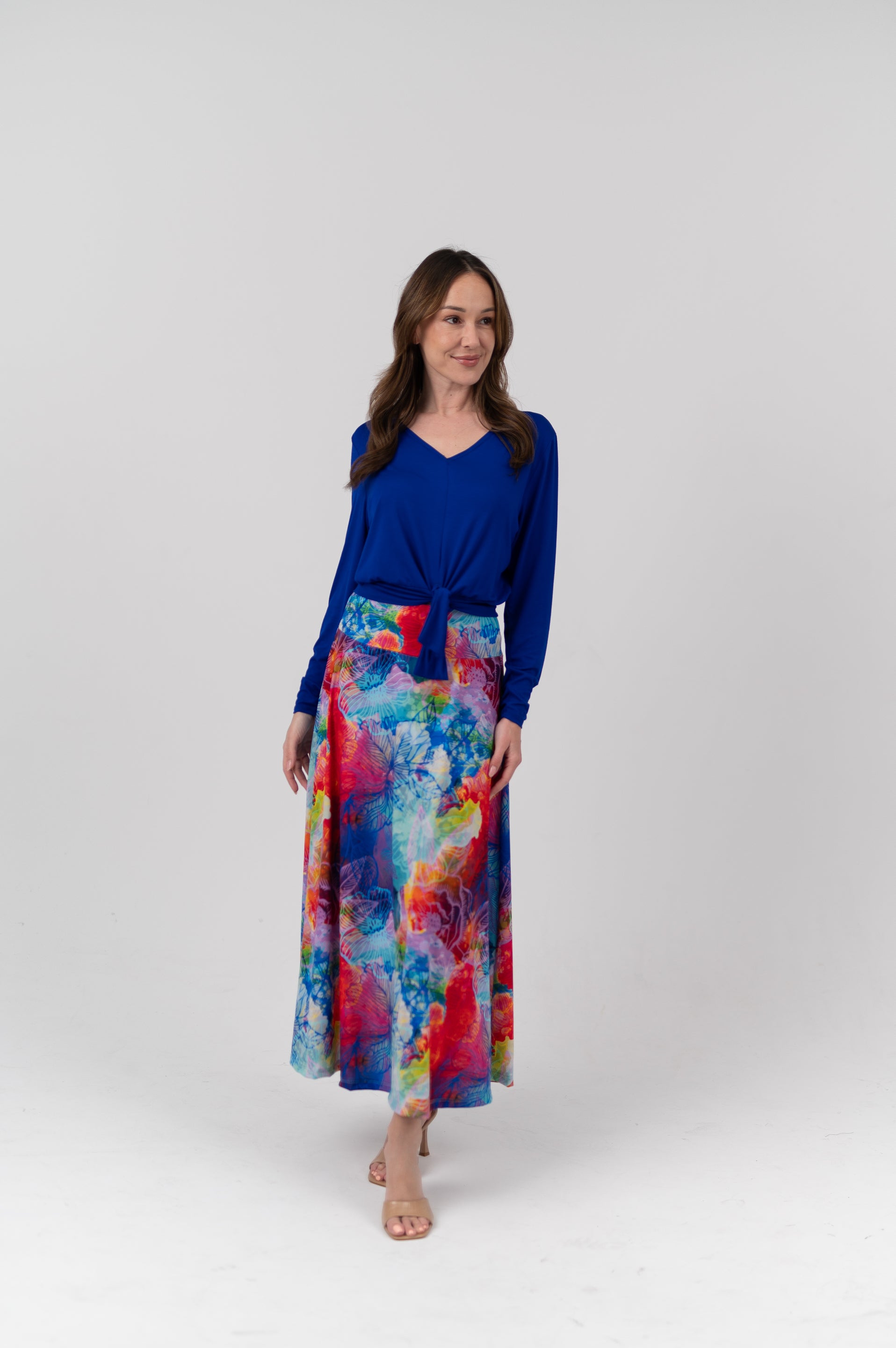 A woman modeling a vibrant, long-sleeved maxi skirt with a colorful abstract watercolor print in shades of blue, pink, red, and yellow, paired with a solid royal blue long-sleeve top, against a light gray studio background.