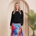 A woman stands in a studio wearing a black long-sleeve top and a colorful floral midi skirt with blue, pink, and purple tones. She is posed in front of a beige arched backdrop with a potted palm plant to the right.