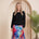 A woman stands in a studio wearing a black long-sleeve top and a colorful floral midi skirt with blue, pink, and purple tones. She is posed in front of a beige arched backdrop with a potted palm plant to the right.
