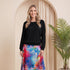 A woman stands in a studio wearing a black long-sleeve top and a colorful floral midi skirt with blue, pink, and purple tones. She is posed in front of a beige arched backdrop with a potted palm plant to the right.