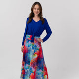 A woman modeling a long-sleeve wrap-style maxi dress with a vibrant blue bodice and a multicolor abstract watercolor print skirt, standing against a light gray studio background.