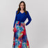 A woman modeling a long-sleeve wrap-style maxi dress with a vibrant blue bodice and a multicolor abstract watercolor print skirt, standing against a light gray studio background.