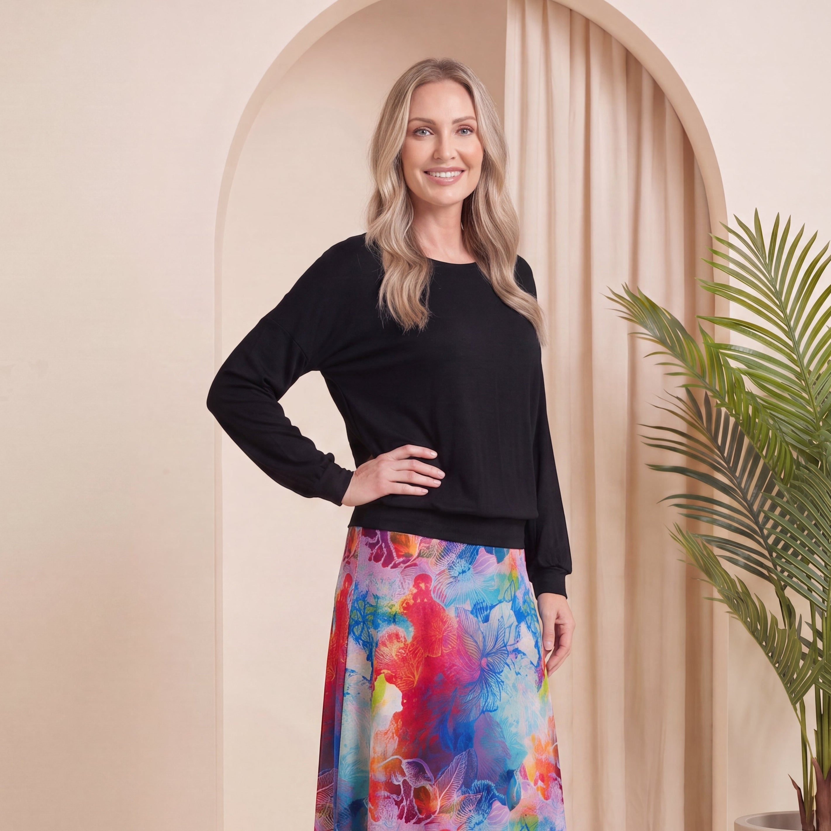 A woman stands in a studio setting wearing a black long-sleeve top and a colorful, watercolor-style midi skirt with beige heeled boots. The background features a beige archway with a draped curtain and a potted palm plant.
