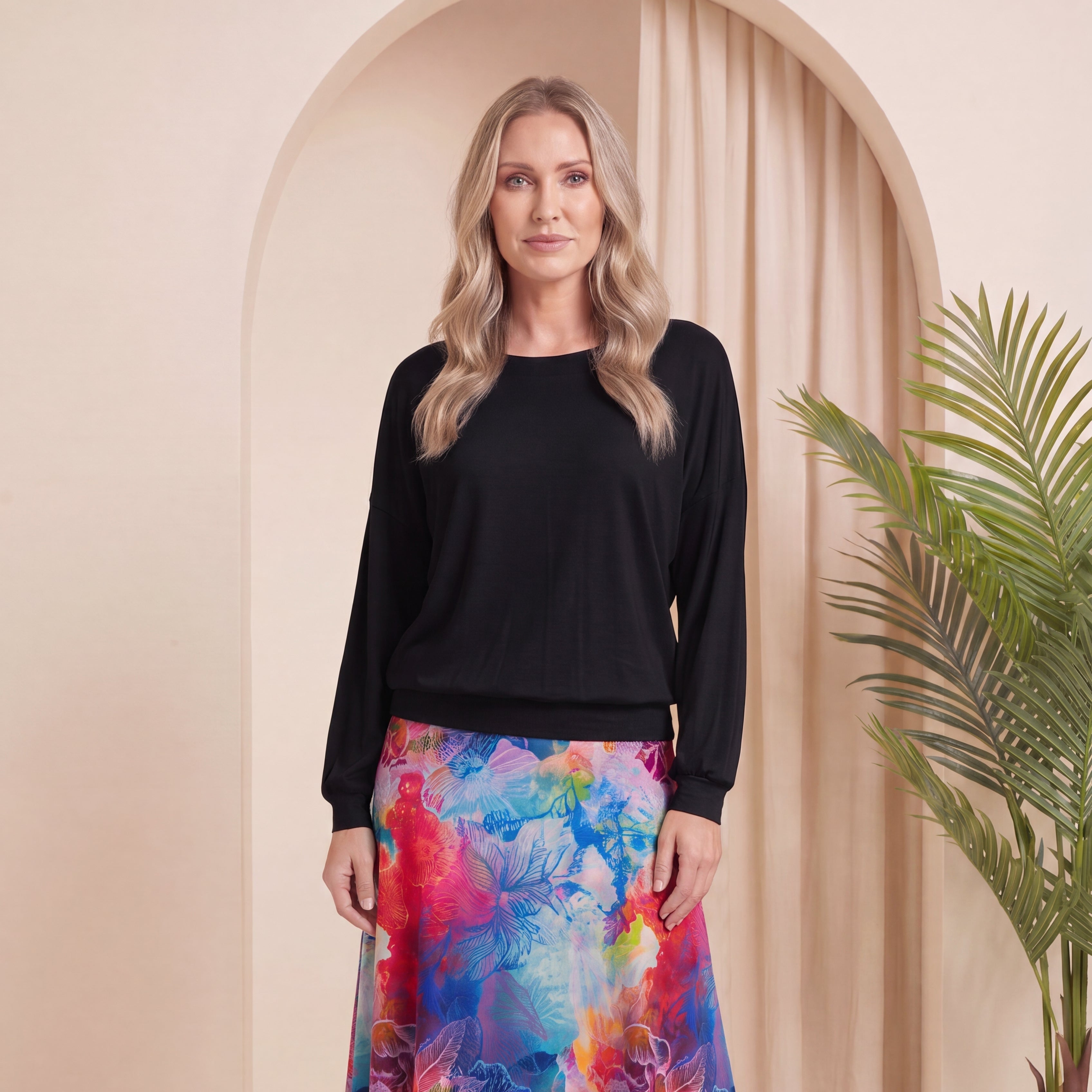 A woman stands in a studio setting wearing a black long-sleeve top and a colorful floral midi skirt with blue, pink, and purple tones, paired with beige ankle boots. An arched beige backdrop and a potted plant are visible behind her.