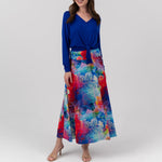 A woman modeling a long-sleeve wrap-style top in royal blue paired with a high-waisted, flared maxi skirt featuring a vibrant abstract watercolor print in red, blue, and pink, set against a plain light gray studio background.
