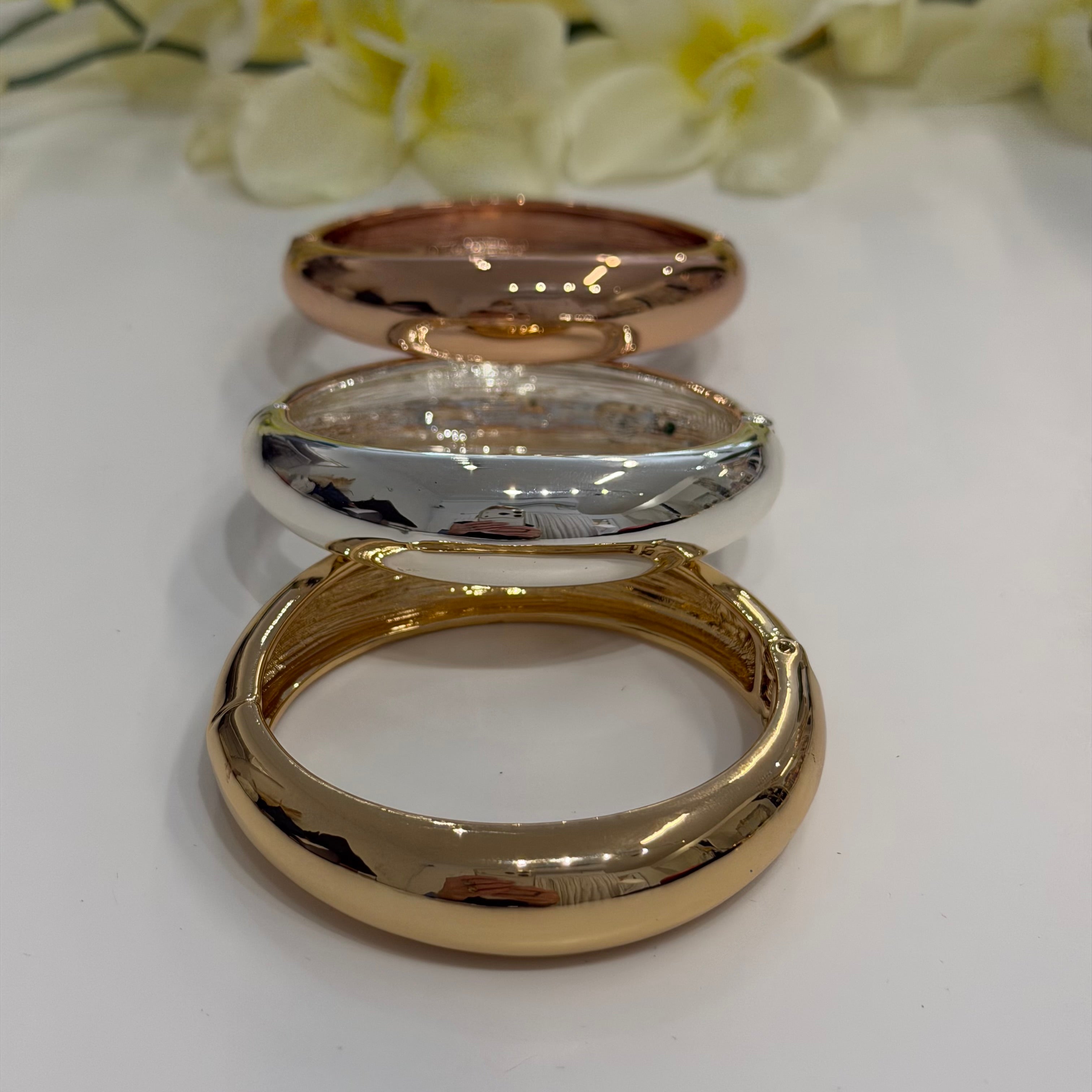 Three gold bangles of different designs on a white surface with flowers in the background.