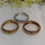 Three bracelets in gold, silver, and rose gold on a white surface with flowers in the background.
