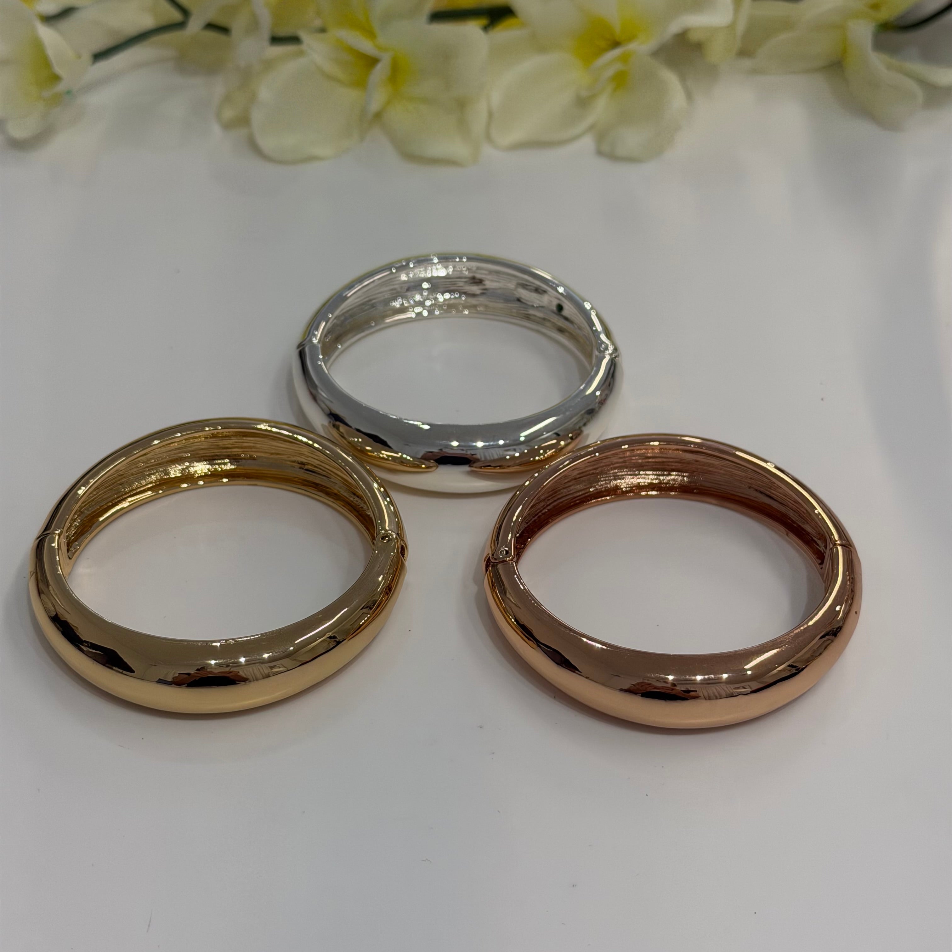 Three bracelets in gold, silver, and rose gold on a white surface with flowers in the background.