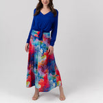 A woman modeling a long-sleeve V-neck top in royal blue paired with a high-waisted, full-length skirt featuring a vibrant multicolor watercolor print in shades of blue, red, pink, and yellow, set against a plain light gray studio background.