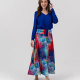 A woman modeling a two-piece outfit consisting of a royal blue long-sleeve V-neck top and a high-waisted, wide-leg maxi skirt with a vibrant multicolor abstract watercolor print in shades of blue, red, pink, and green, set against a plain light gray studio background.