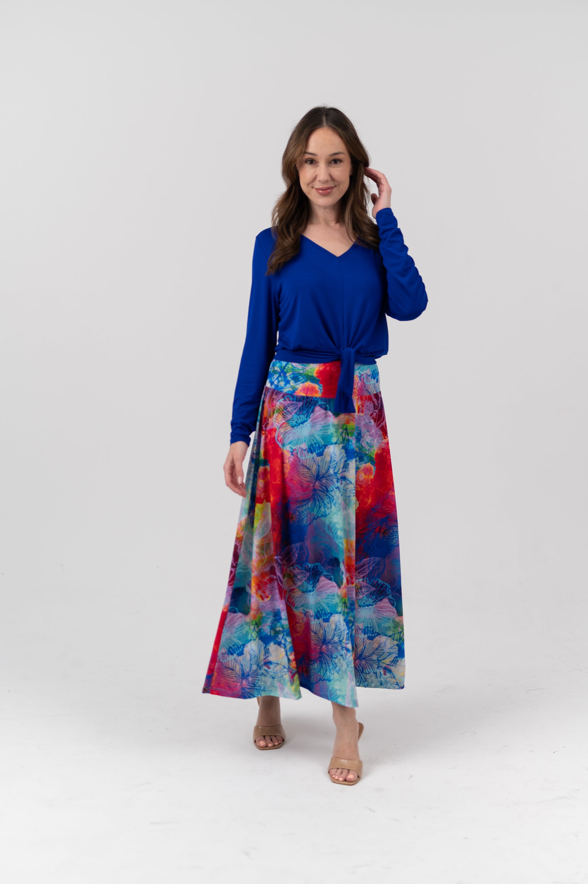 A woman modeling a two-piece outfit consisting of a royal blue long-sleeve V-neck top and a high-waisted, wide-leg maxi skirt with a vibrant multicolor abstract watercolor print in shades of blue, red, pink, and green, set against a plain light gray studio background.