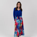 A woman modeling a long-sleeve royal blue V-neck top paired with a high-waisted, wide-leg maxi skirt featuring a vibrant multicolor abstract watercolor print in shades of blue, red, pink, and yellow, set against a plain light gray studio background.