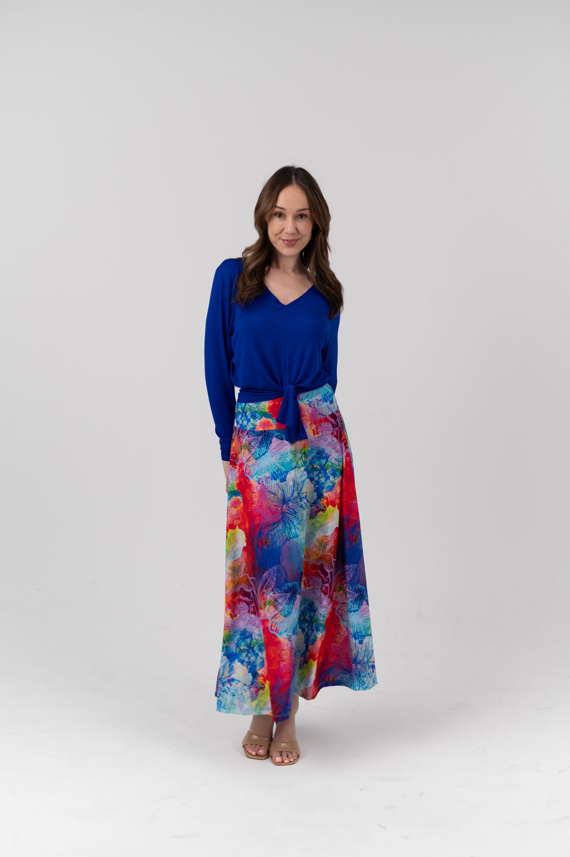 A woman modeling a long-sleeve royal blue V-neck top paired with a high-waisted, wide-leg maxi skirt featuring a vibrant multicolor abstract watercolor print in shades of blue, red, pink, and yellow, set against a plain light gray studio background.