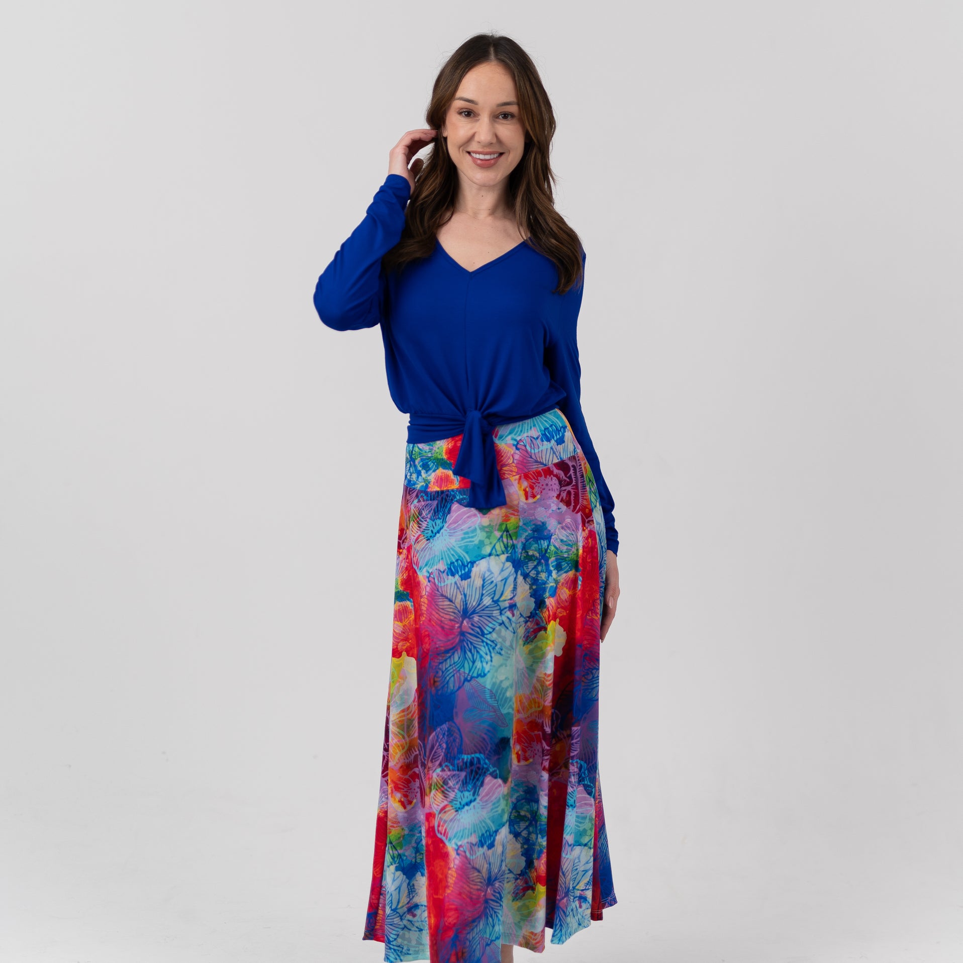 A woman modeling a long-sleeve V-neck top in royal blue paired with a high-waisted, ankle-length skirt featuring a vibrant multicolor abstract watercolor print in shades of blue, red, pink, and yellow, set against a plain light gray studio background.