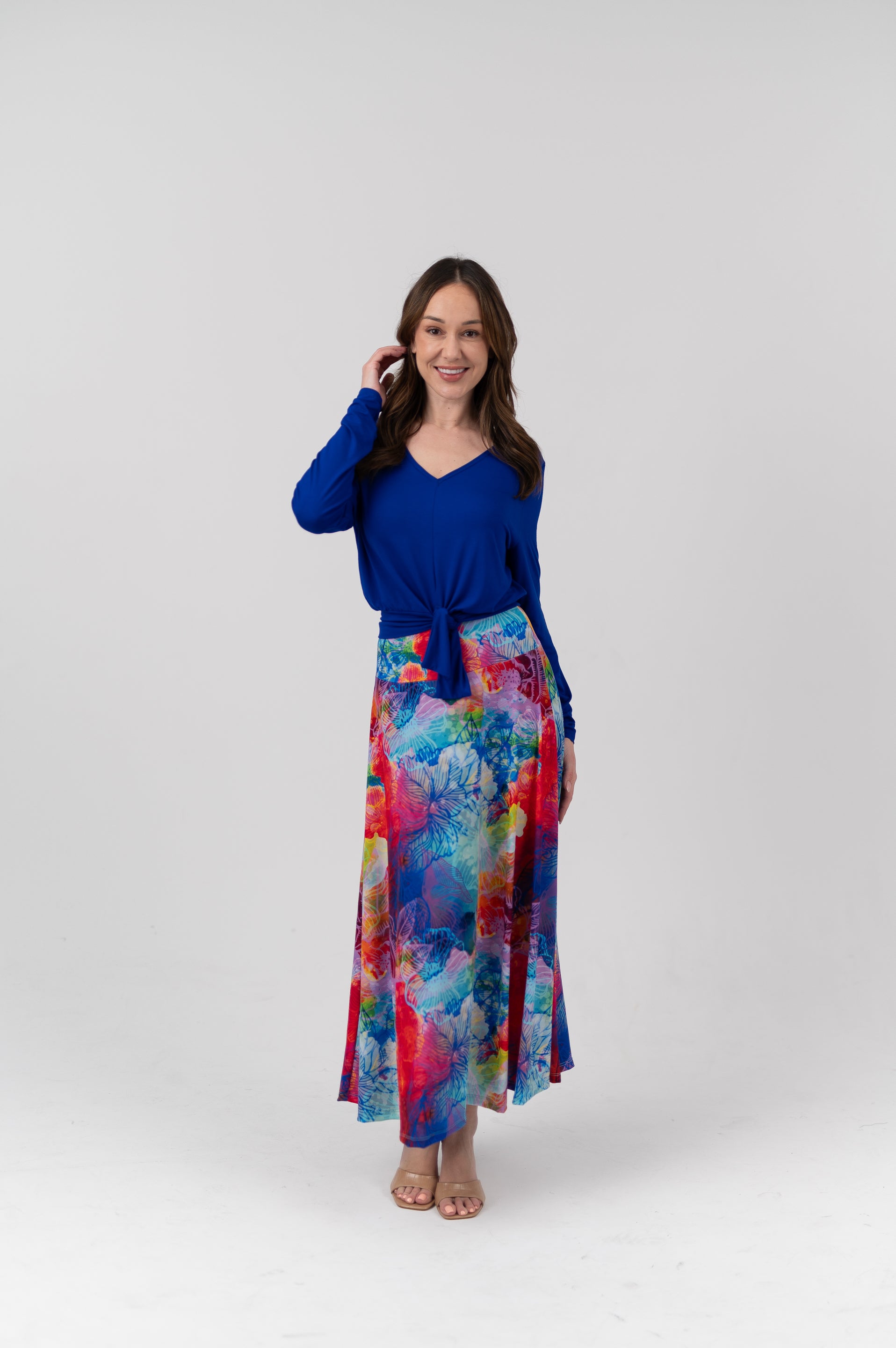 A woman modeling a long-sleeve V-neck top in royal blue paired with a high-waisted, ankle-length skirt featuring a vibrant multicolor abstract watercolor print in shades of blue, red, pink, and yellow, set against a plain light gray studio background.