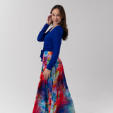 A woman modeling a vibrant, multicolored abstract-print maxi skirt paired with a royal blue long-sleeve top, standing against a plain light gray studio background.